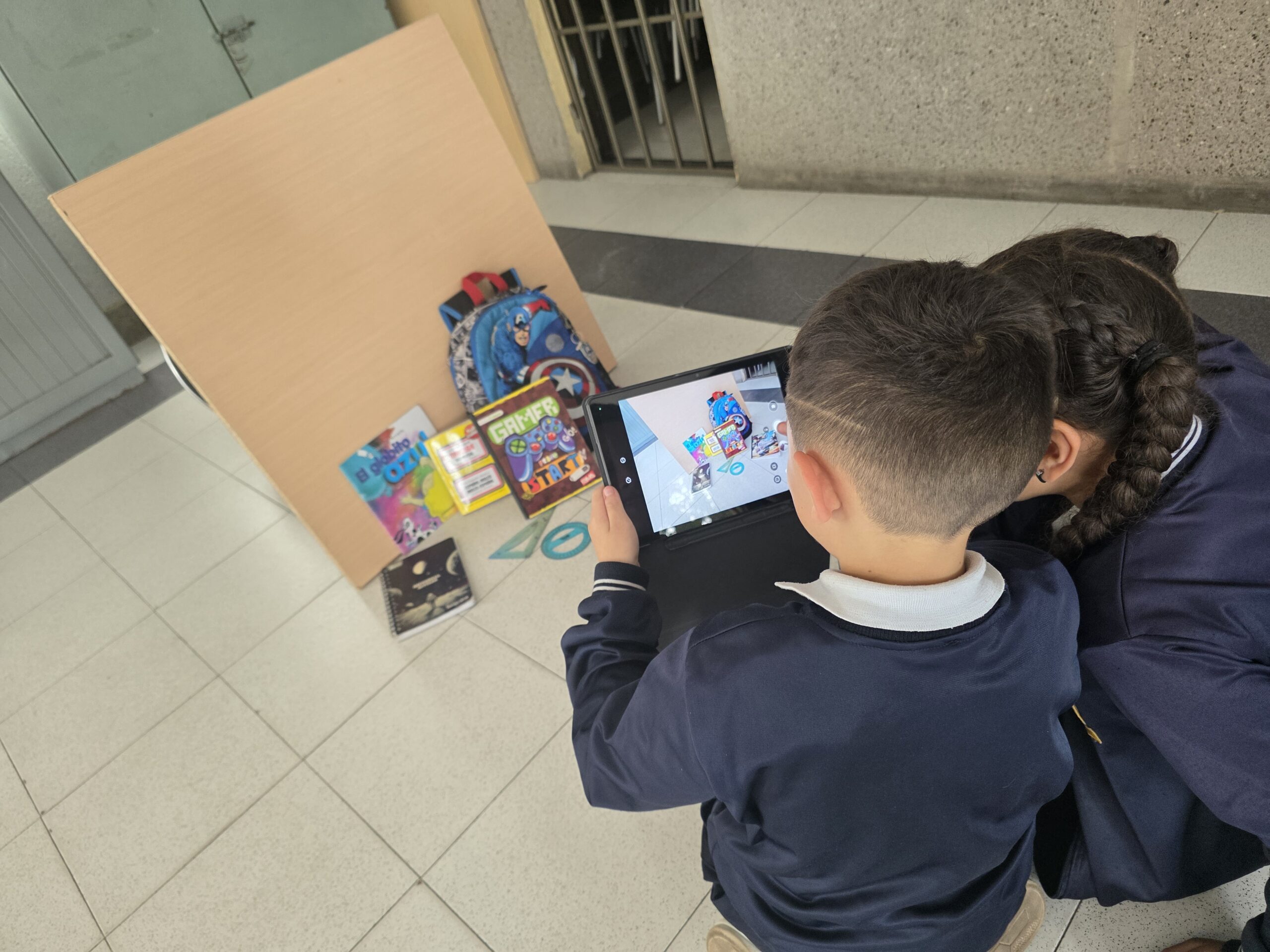 Dos estudiantes fotografían con una tablet una composición de libros y morral sobre el piso