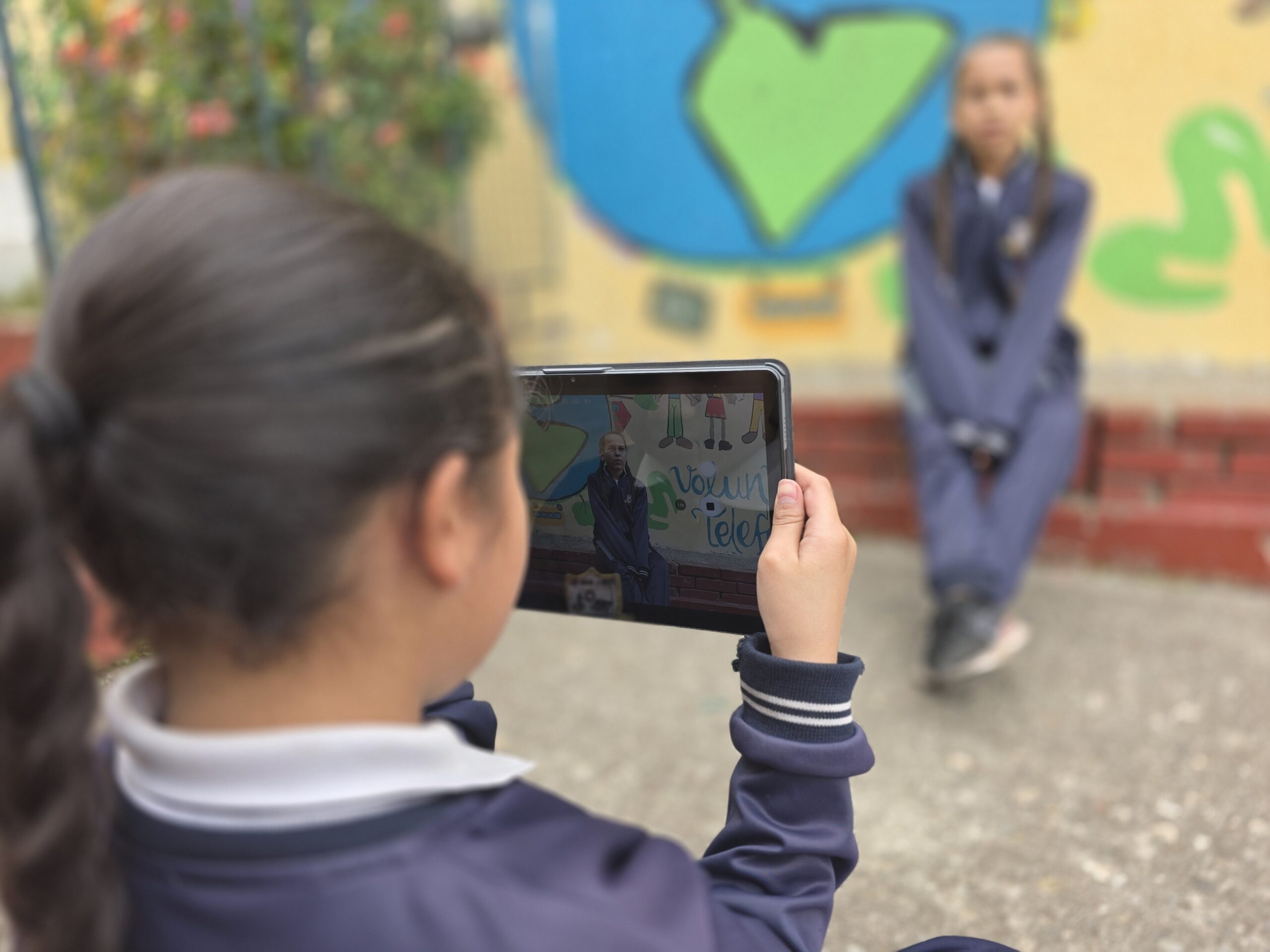 Estudiante fotografía con la tablet a una compañera sentada frente a un mural del planeta