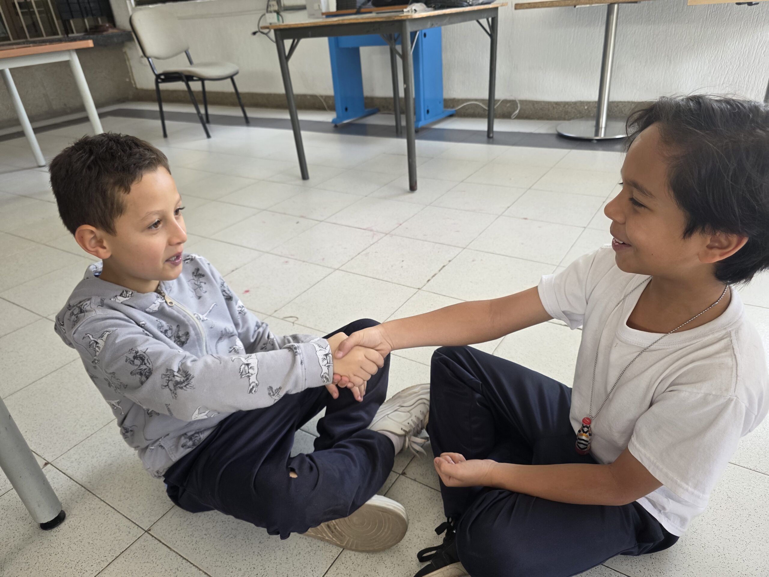 Dos estudiantes sentados en el piso se dan la mano y sonríen durante el juego de manos del taller