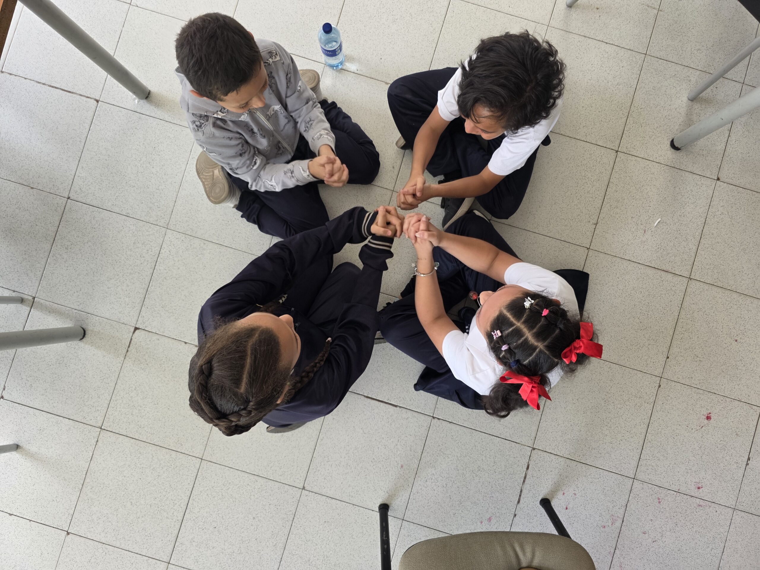 Vista cenital de cuatro estudiantes sentados en círculo cuyas manos se encuentran en el centro durante el juego