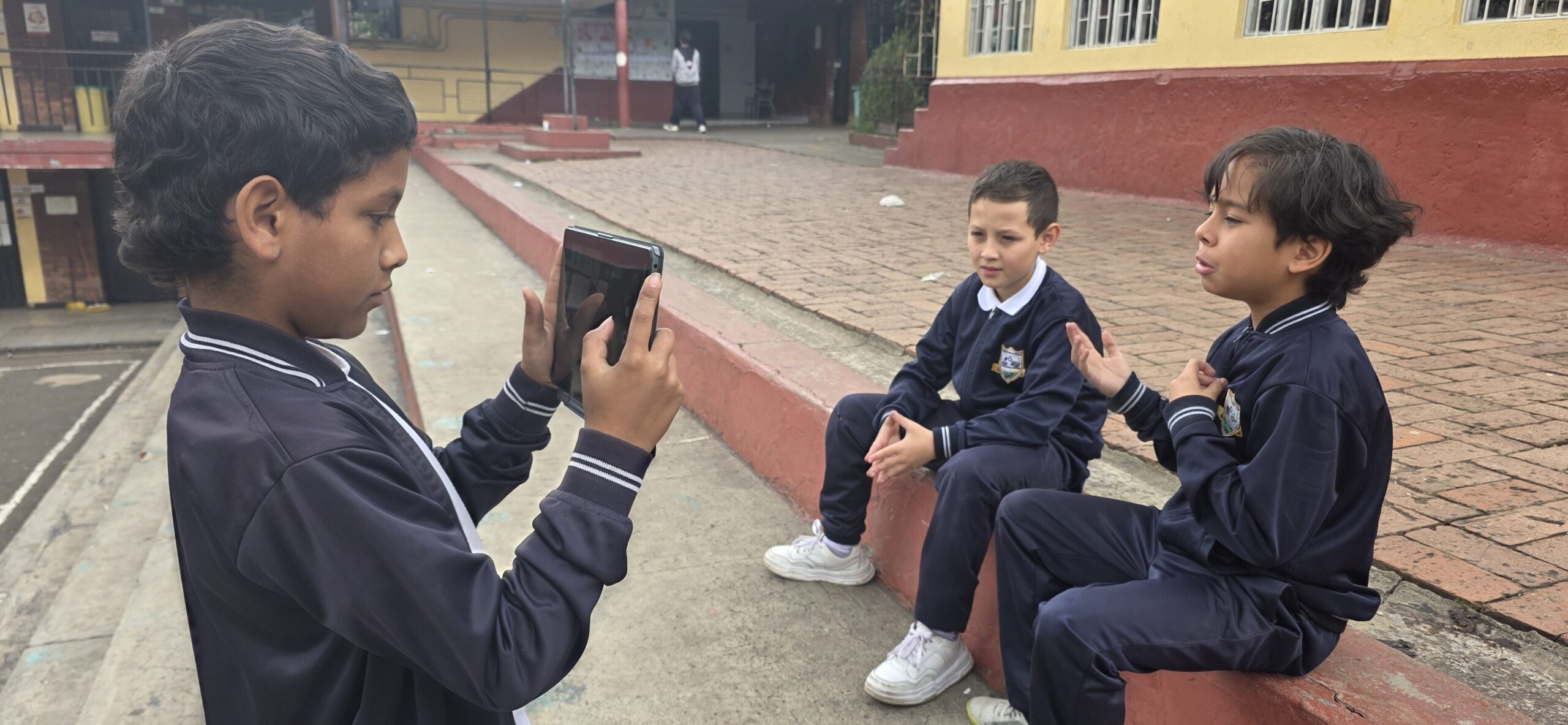 Un estudiante graba con una tablet a dos compañeros sentados en el borde de la jardinera del patio