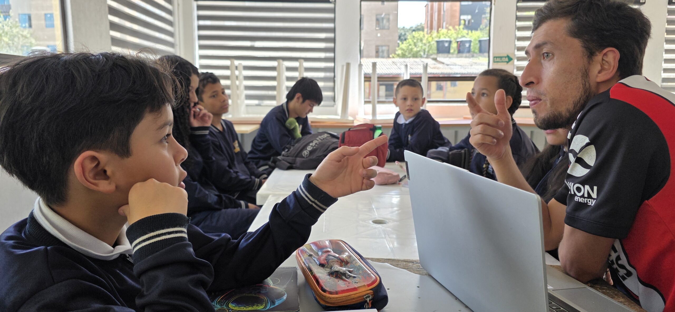 El docente del taller explica con las manos cómo preparar una entrevista mientras los estudiantes escuchan alrededor de la mesa