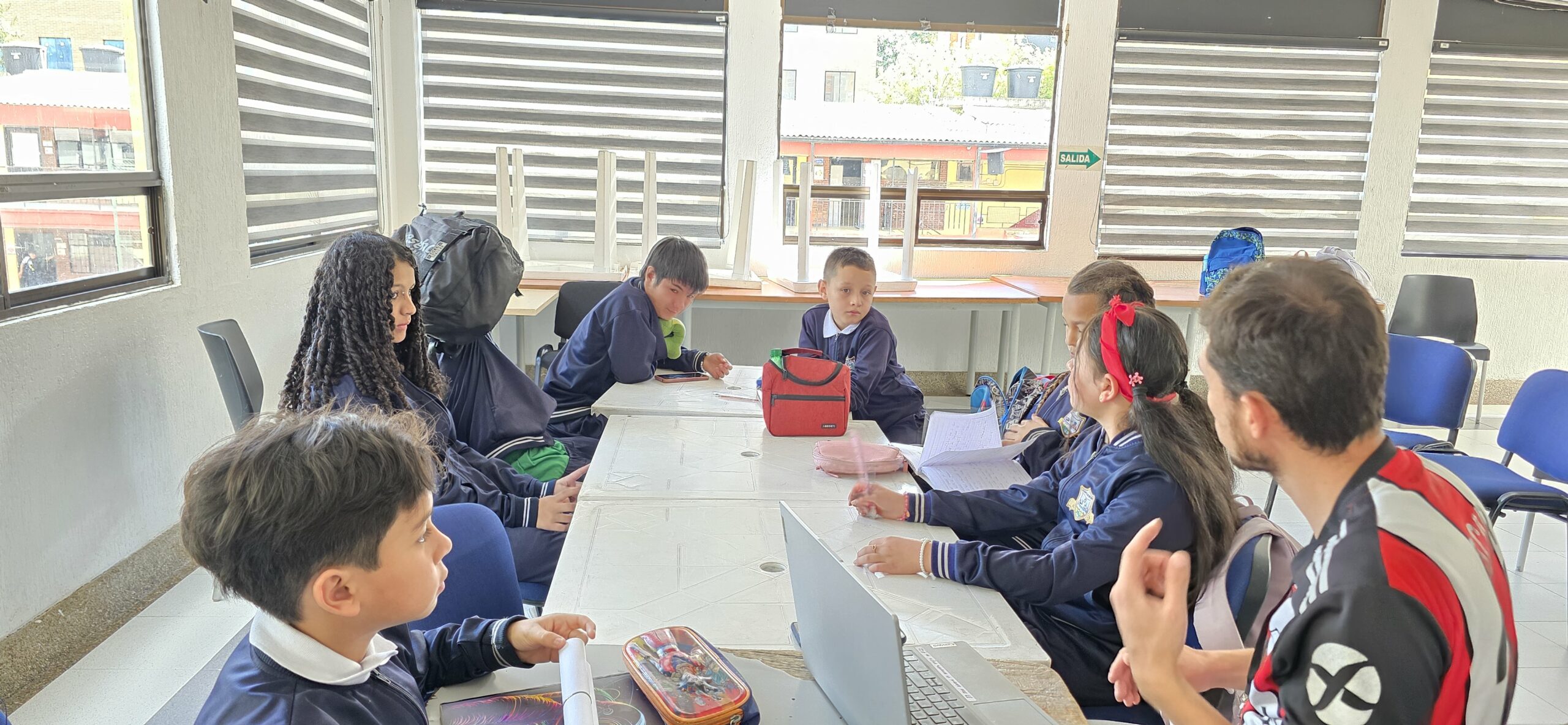 Estudiantes escriben sus guiones de entrevista en cuadernos mientras trabajan con tablets y portátiles en el salón