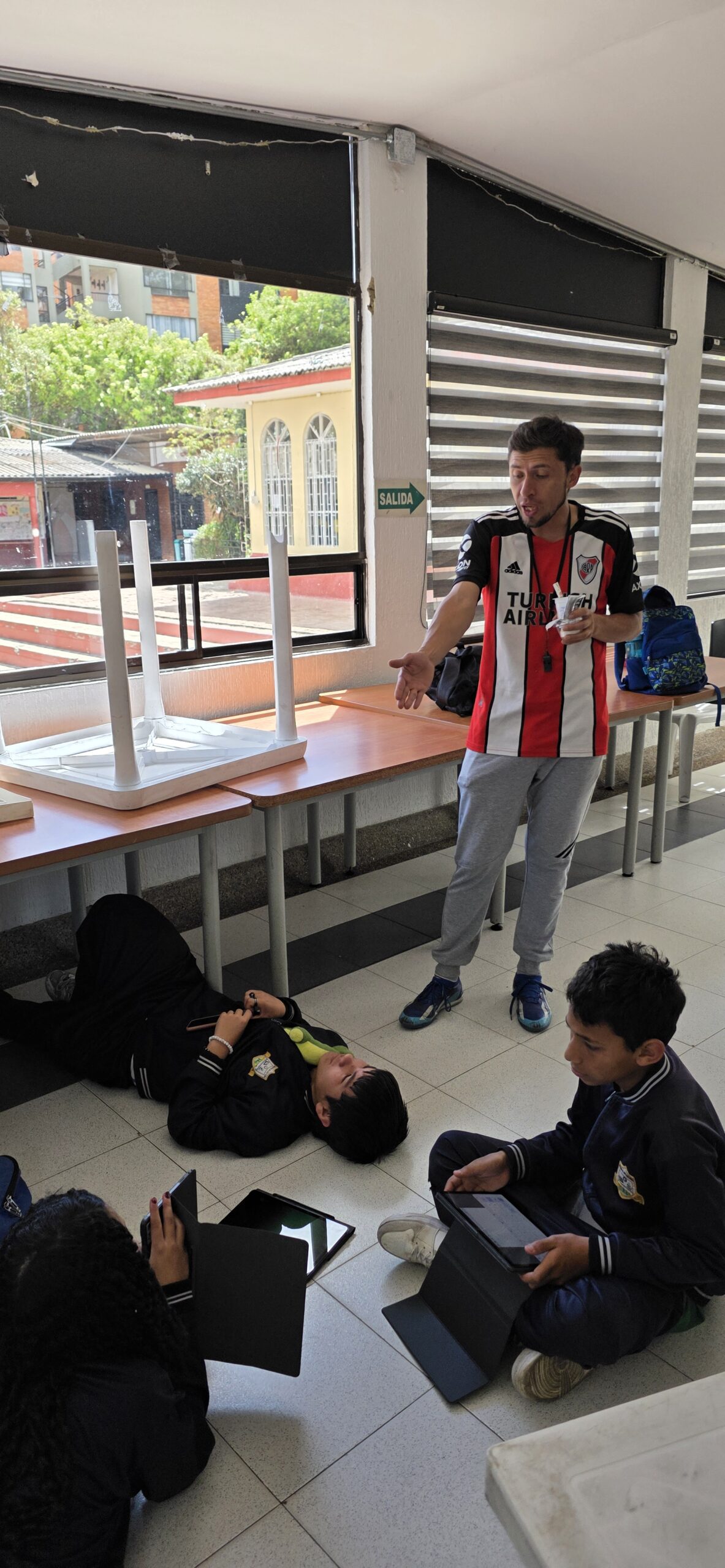 Estudiantes sentados en el piso del salón revisan sus tablets mientras el docente acompaña de pie