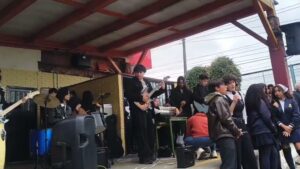 Estudiantes del coro cantan con micrófono durante la presentación en la plazoleta, con la banda al fondo
