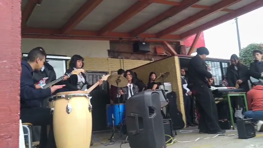 El semillero musical toca en vivo en la plazoleta cubierta del colegio, con tumbadoras y guitarra en primer plano