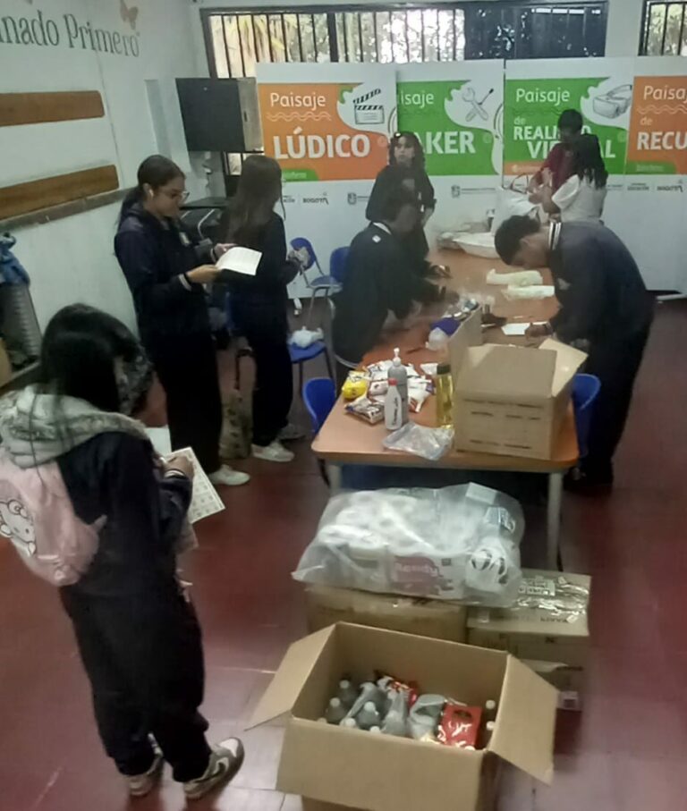 Estudiantes clasifican y empacan las donaciones sobre las mesas de un salón, con cajas y paquetes de papel en el piso