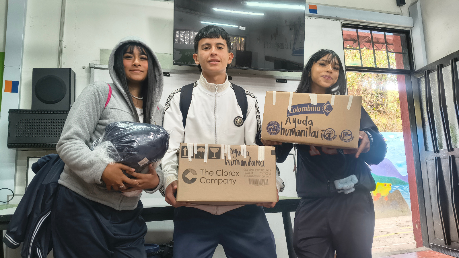 Tres estudiantes sostienen en un salón dos cajas marcadas a mano con «Ayuda humanitaria» y una cobija empacada
